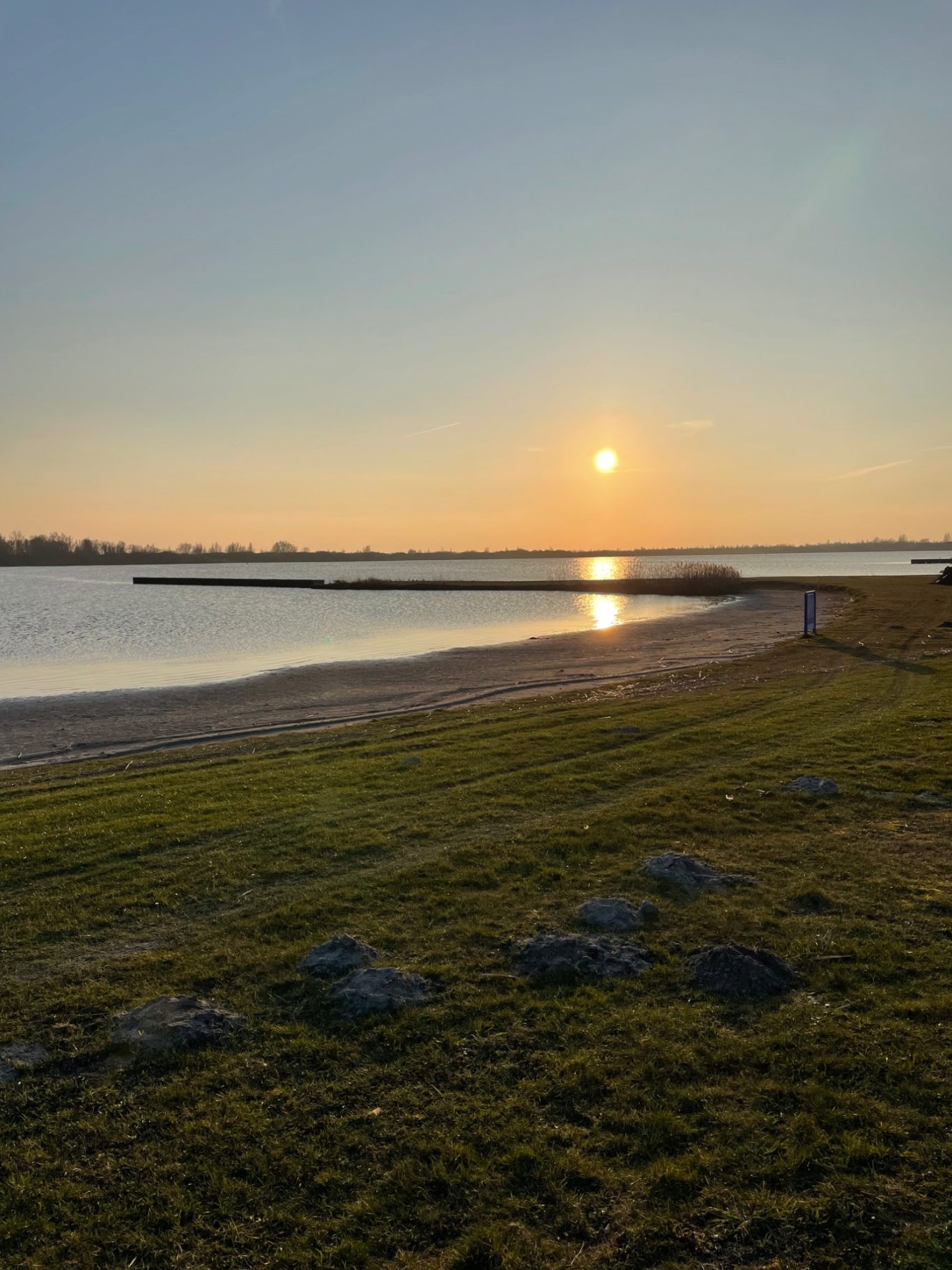 Badestrand Sonnenuntergang über einem ruhigen Gewässer mit Strand und Steg im Vordergrund.