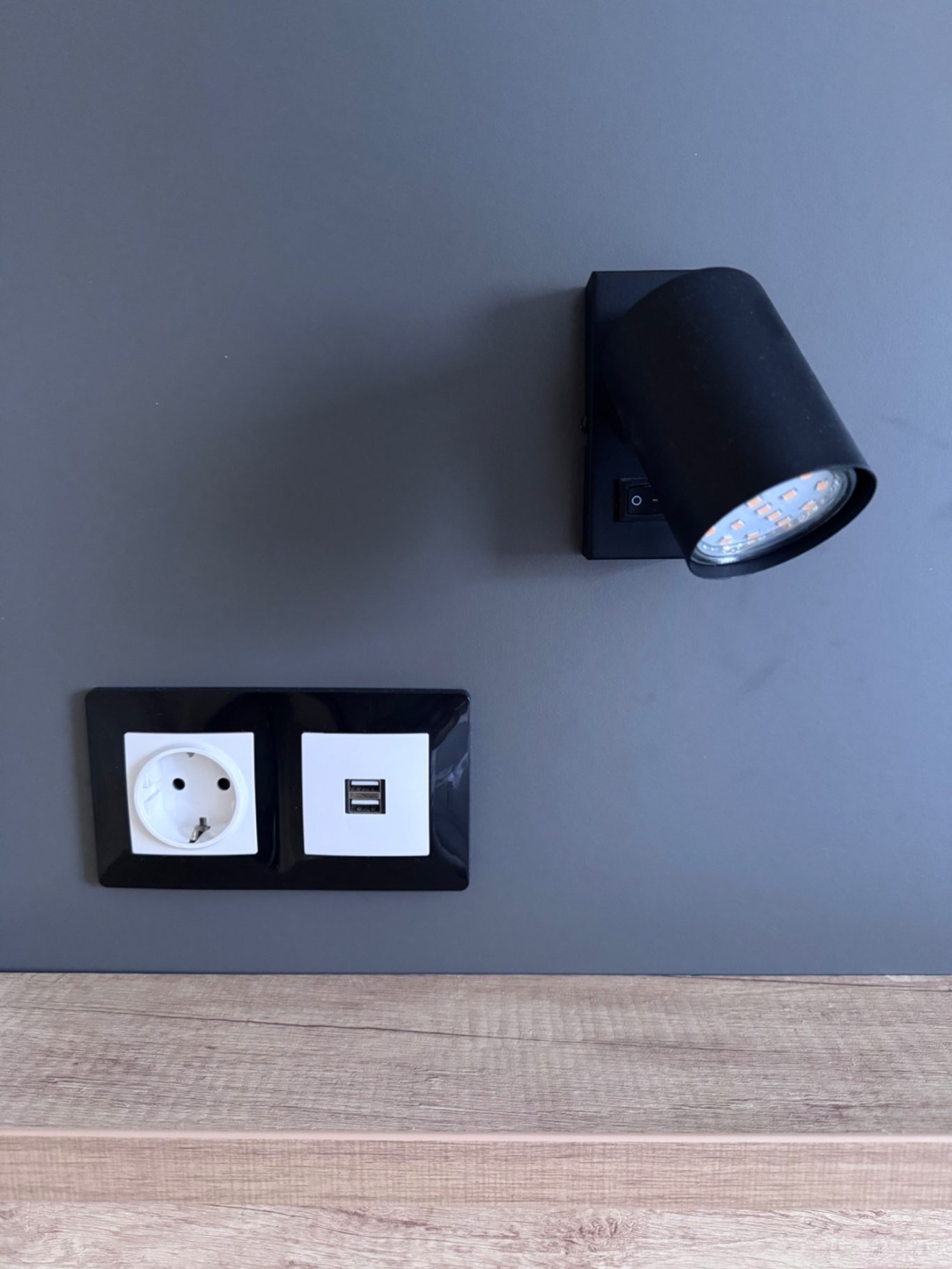Einrichtungsdetailles Schwarze Wandlampe neben einer Steckdose und einem USB-Anschluss auf grauer Wand.