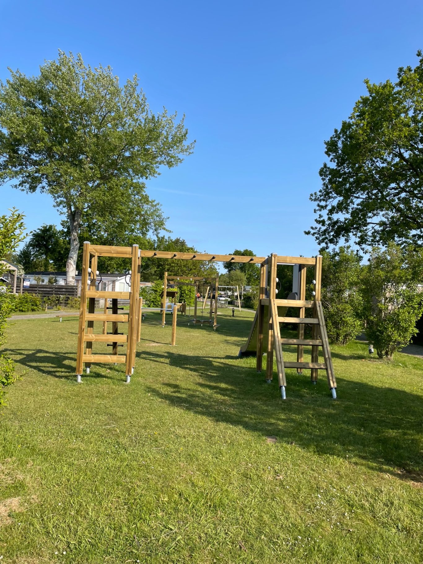 Spielplätze Holzspielplatz mit Schaukeln und Rutsche, umgeben von Rasen und Bäumen.
