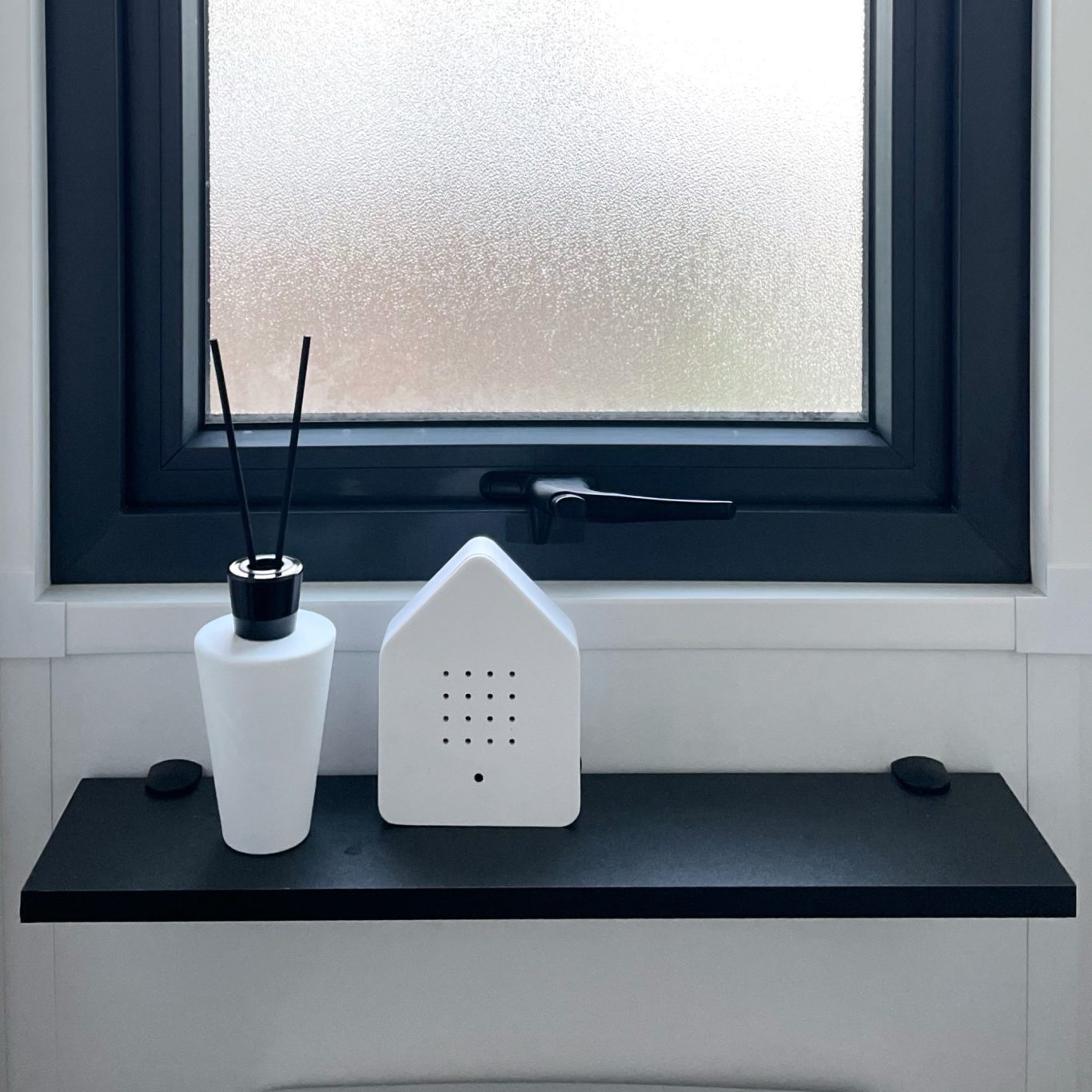 Einrichtungsdetailles Weißer Diffusor und dekoratives Haus auf einem schwarzen Regal vor einem Fenster.