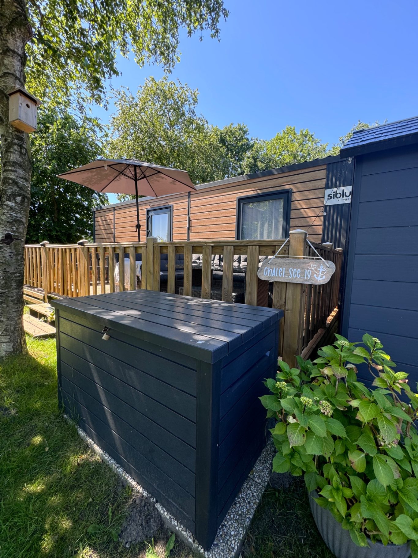 Aussenansicht Hölzerne Terrasse mit Sonnenschirm neben einem Gartenhäuschen und bepflanztem Bereich.